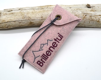 May include: A pink felt glasses case with a mountain graphic and the word "Brillenetui" embroidered in dark red. The case has a flap closure with a dark gray cord and a metal grommet.