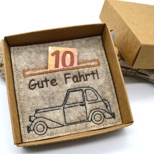 May include: A tan cardboard gift box with a felt insert. The insert features an embroidered vintage car and the German phrase "Gute Fahrt!" with a ten-dollar bill. The box is open, revealing the contents.