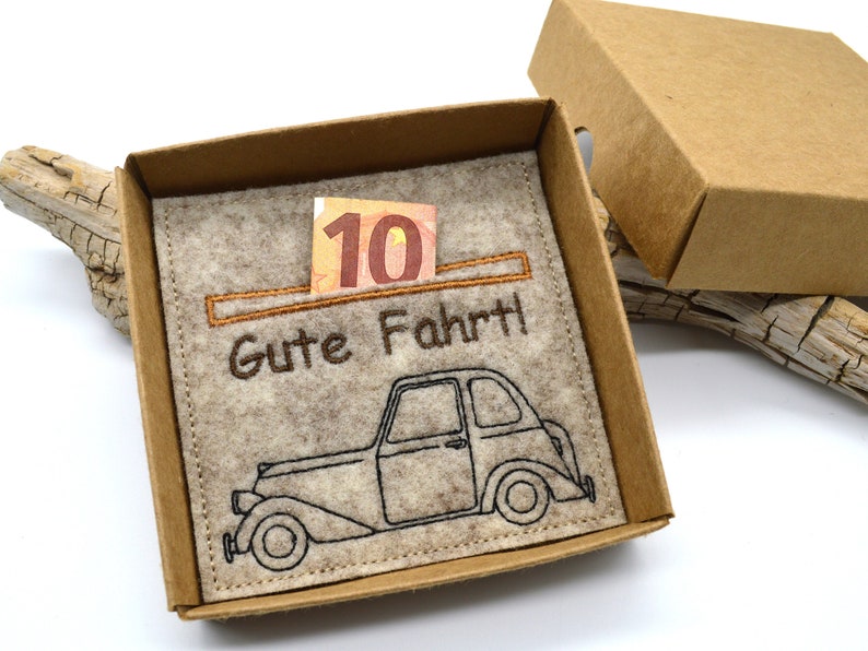 May include: A tan cardboard gift box with a felt insert. The insert features an embroidered vintage car and the German phrase "Gute Fahrt!" with a ten-dollar bill. The box is open, revealing the contents.
