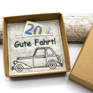 May include: A gift box with a felt insert featuring a vintage car illustration and the German phrase "Gute Fahrt!" (Safe travels!). A twenty-euro note is visible in the pocket. The box is made of brown cardboard.