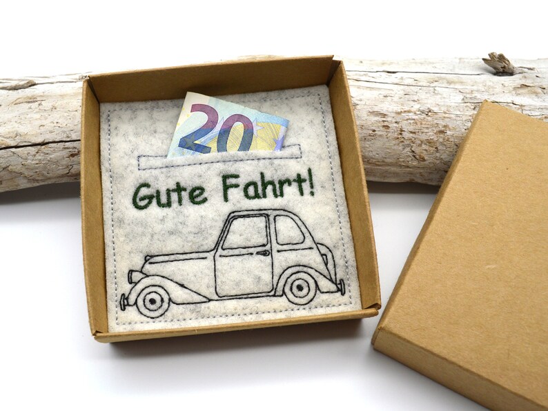May include: A gift box with a felt insert featuring a vintage car illustration and the German phrase "Gute Fahrt!" (Safe travels!). A twenty-euro note is visible in the pocket. The box is made of brown cardboard.