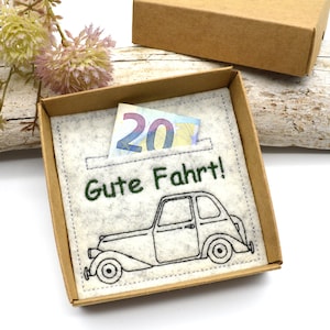 May include: A gift box with a felt insert featuring an embroidered vintage car and the phrase "Gute Fahrt!" in green. A twenty-euro bill is visible in a pocket. The box is brown.