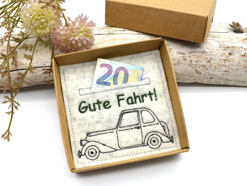 May include: A gift box with a felt insert featuring an embroidered vintage car and the phrase "Gute Fahrt!" in green. A twenty-euro bill is visible in a pocket. The box is brown.