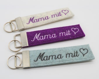 May include: Three rectangular keychains in cream, purple, and teal. Each keychain has the embroidered text "Mama mit" with a heart symbol. The keychains have silver-colored metal hardware.