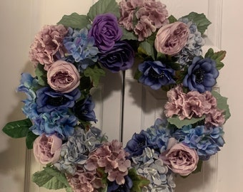 May include: A purple, blue and pink floral wreath featuring hydrangeas, roses and anemones. The wreath is made of artificial flowers and is ideal for decorating a door or wall.