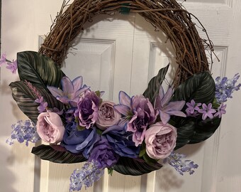 May include: A grapevine wreath decorated with purple and pink flowers, including roses and lilies, with green leaves.