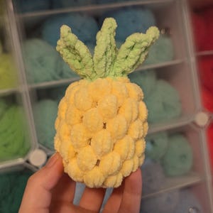 May include: A handmade, plush pineapple toy. The pineapple is yellow with a textured surface, and the top has green leaves. The toy is held in a person's hand, with a background of colorful yarn.