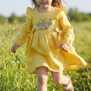 May include: A young child wearing a yellow dress with long sleeves and a bunny and floral embroidery on the bodice. The dress has a ruffled neckline and is paired with white socks and brown sandals. The child is running through a field of tall green grass.