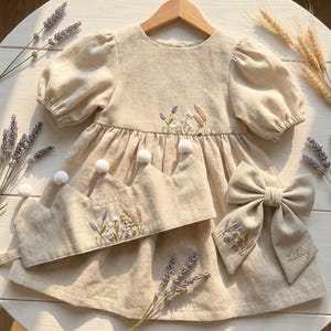 May include: A beige dress with puff sleeves, a matching crown with white pom-poms, and a hair bow. The dress, crown, and bow feature embroidered floral designs and wheat stalks. The name Lilly is embroidered on the crown and the hair bow.