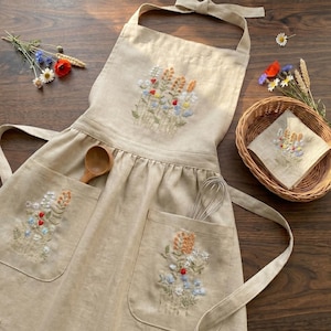 Könnte beinhalten: Eine beigefarbene Schürze mit gestickten Wildblumensträußen auf der Brust und zwei Vordertaschen. Die Schürze hat passende Stoffbänder und wird von einem Weidenkorb begleitet, der eine quadratische Stoffserviette mit gleichem Blumenmuster enthält.