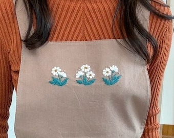 May include: A tan apron with embroidered white flowers and green leaves. The apron has a tie waist and two front pockets. The model is wearing a rust-colored turtleneck sweater underneath the apron.
