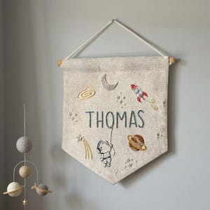 Könnte beinhalten: Ein beiges Banner mit dem Namen "THOMAS" in Dunkelblau gestickt, mit Weltraum-Motiven wie einer Rakete, Planeten und einem Astronauten. Das Banner hängt über einem Holzbettchen, mit einem Mobile und einem Buch mit einer Rakete.