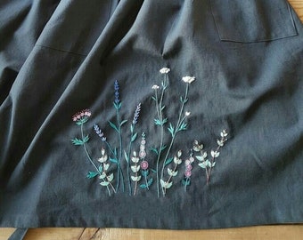 May include: A dark gray apron with two front pockets and a floral embroidery design. The embroidery features various wildflowers in shades of pink, purple, white, and green. The apron has a gathered waist and long ties.
