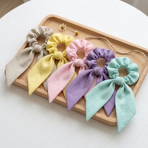 May include: A set of five fabric hair scrunchies with long ribbon tails, arranged in a row. The colors include beige, yellow, light pink, lavender, and mint green. Each scrunchie features a gathered elastic band and two dangling fabric strips.