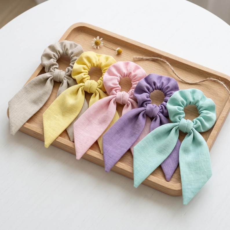 May include: A set of five fabric hair scrunchies with long ribbon tails, arranged in a row. The colors include beige, yellow, light pink, lavender, and mint green. Each scrunchie features a gathered elastic band and two dangling fabric strips.