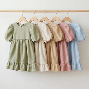 May include: Five dresses hanging on wooden hangers, each featuring short puff sleeves, a round neckline, a gathered waist, and a ruffled hem. The dresses are displayed in a row in shades of sage green, cream, tan, dusty rose, and light blue.