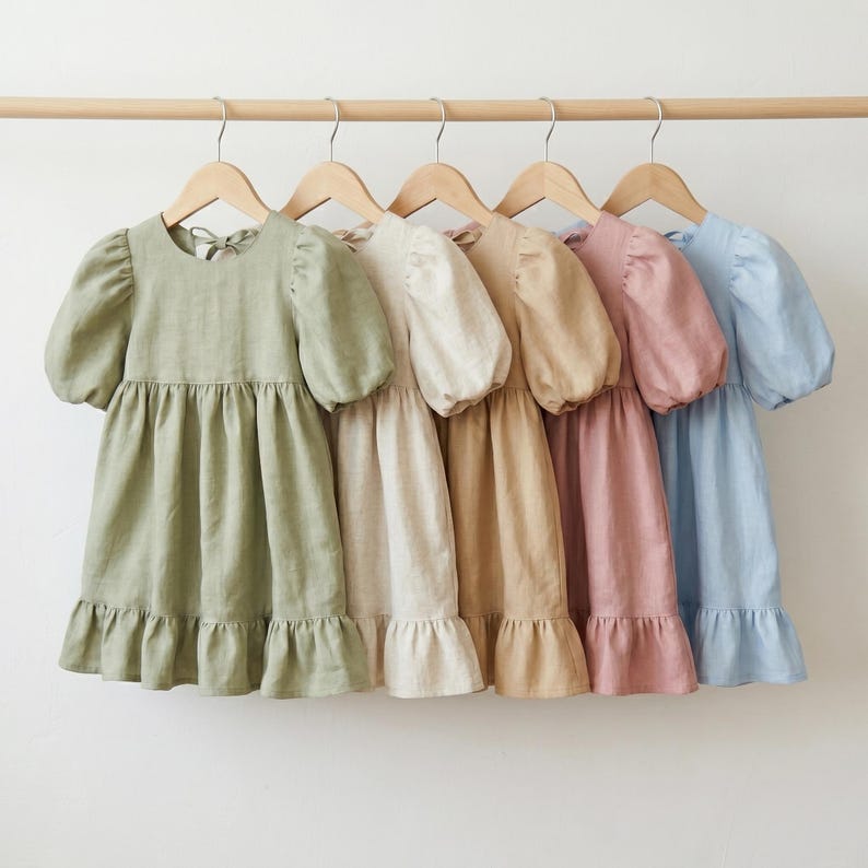 May include: Five dresses hanging on wooden hangers, each featuring short puff sleeves, a round neckline, a gathered waist, and a ruffled hem. The dresses are displayed in a row in shades of sage green, cream, tan, dusty rose, and light blue.