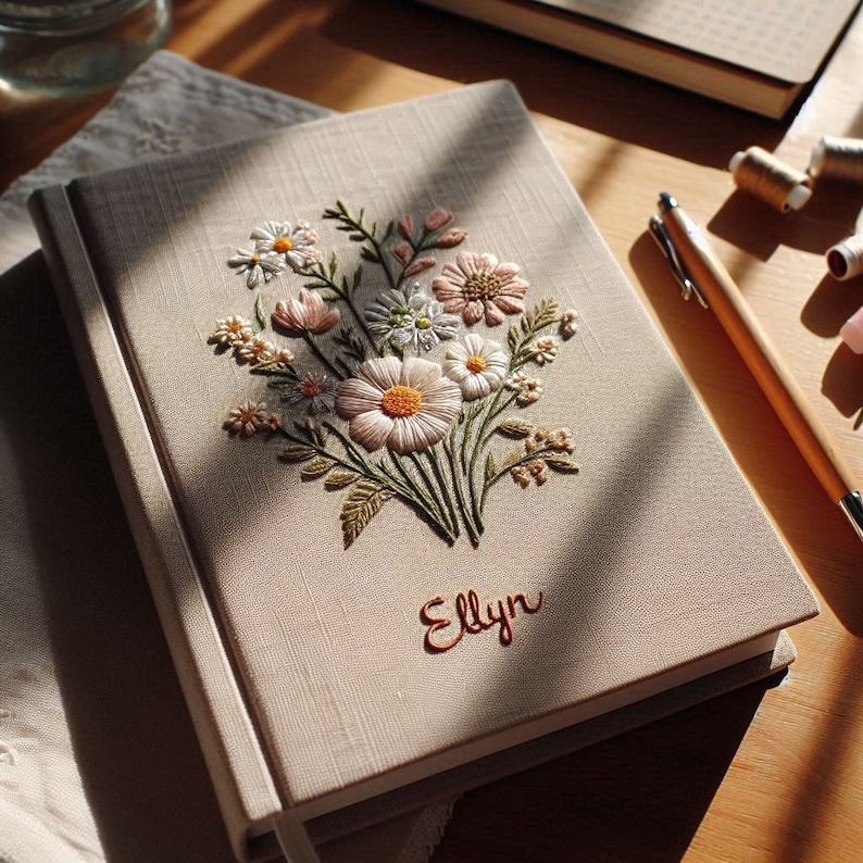 Op de afbeelding: Een beige linnen dagboek met een geborduurd bloemenmotief met witte en roze bloemen. De naam "Elly" is geborduurd in bruin garen op de voorkant.
