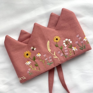 Peut inclure: Une couronne en tissu rose avec des fleurs, des feuilles et une abeille brodées. La couronne a une longue attache en tissu rose. Le mot "Nora" est brodé sur la couronne.