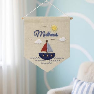 Op de afbeelding: Geborduurde wanddecoratie met een nautisch thema. Voorzien van een zeilboot met een rood en blauw zeil, blauwe golven en de naam "Matheus" in blauw schrift. Inclusief een gele zon en witte wolken.