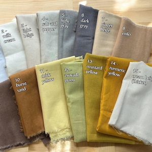 Op de afbeelding: Een verzameling van 15 stofstalen in verschillende tinten beige, bruin, geel, groen en grijs. De stalen zijn gelabeld met nummers en namen, zoals "zuiver wit", "licht beige", "gebrand zand" en "avocado groen".