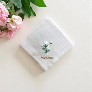 May include: A white linen handkerchief with a small embroidered flower and bee design. The handkerchief has the date "15.01.2025" embroidered in brown thread.