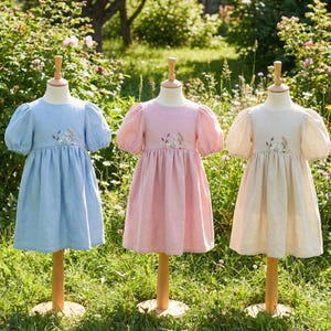 May include: Three dresses displayed on mannequins, each featuring puff sleeves, a round neckline, and a gathered waist. The dresses are light blue, soft pink, and beige, with delicate floral embroidery centred on the chest of each garment.