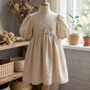 May include: A beige linen dress featuring short puff sleeves, a round neckline, and a gathered skirt. The bodice is decorated with a delicate, embroidered botanical design of small wildflowers and grasses centred at the waistline.
