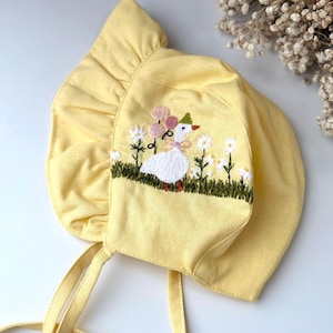 Op de afbeelding: Een gele baby-bonnet met een geborduurde witte eend erop. De eend houdt ballonnen vast en is omringd door madeliefjes. De bonnet heeft twee gele banden.