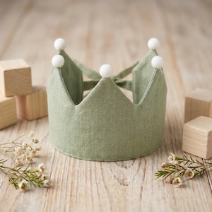 May include: A sage green fabric crown featuring five pointed peaks, each topped with a small white pom-pom. The crown is constructed from a textured linen-like material and includes fabric ties at the back for an adjustable fit around the head.