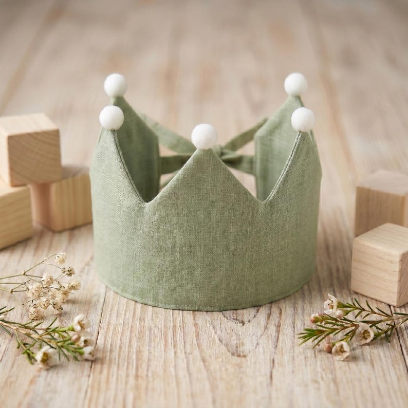 May include: A sage green fabric crown featuring five pointed peaks, each topped with a small white pom-pom. The crown is constructed from a textured linen-like material and includes fabric ties at the back for an adjustable fit around the head.