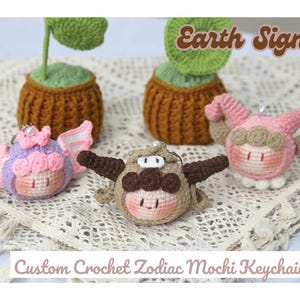 May include: Four crocheted zodiac mochi keychain charms, each with a different earth sign: Taurus, Virgo, and Capricorn. The charms are pink, brown, and purple and are displayed on a crocheted surface with the text "Earth Signs" and "Custom Crochet Zodiac Mochi Keychain".