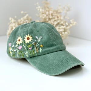 Pode incluir: Um boné de beisebol verde com flores bordadas em tons de rosa, amarelo e verde.