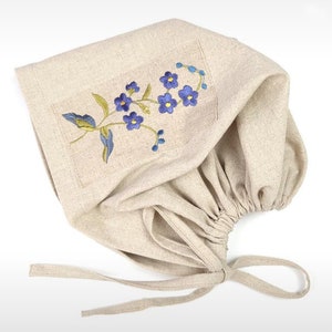 Puede incluir: Un gorro de lino beige con un diseño de bordado floral azul y verde. El gorro tiene un cordón en la parte inferior y lazos en la parte posterior.