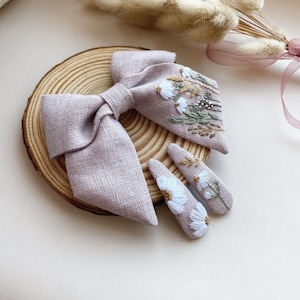 May include: A set of two hair clips with a light purple linen bow and embroidered white flowers. The bow is tied with a knot in the center and the clips are decorated with white flowers and green leaves.