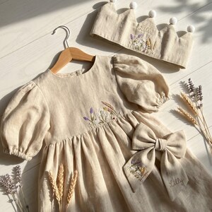 May include: A beige dress with puff sleeves and floral embroidery, a matching crown with white pom-poms and the name Lilly, and a large hair bow featuring floral embroidery and the name Lilly, all crafted from a light-coloured fabric.
