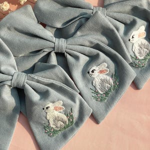 Peut inclure: Trois nœuds en tissu bleu clair avec des lapins blancs brodés. Chaque nœud présente un motif de lapin différent.