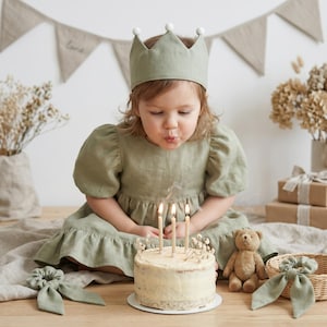 May include: A sage green linen dress with puff sleeves and a ruffled hem, paired with a matching crown featuring white pom-poms. A round cake with three lit candles sits in front, accompanied by a small brown teddy bear and two hair scrunchies.