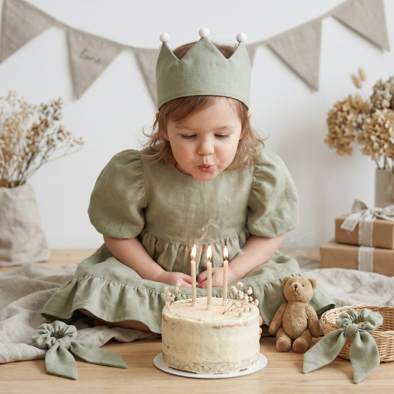 May include: A sage green linen dress with puff sleeves and a ruffled hem, paired with a matching crown featuring white pom-poms. A round cake with three lit candles sits in front, accompanied by a small brown teddy bear and two hair scrunchies.