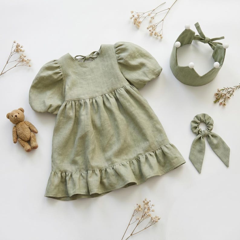 May include: A sage green dress with puff sleeves and a ruffled hem, accompanied by a matching crown headband with white pom-poms and a hair scrunchie with long fabric tails. A small brown teddy bear is positioned to the left of the dress.