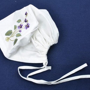 Puede incluir: Bonnet de algodón blanco con un diseño de bordado de flores moradas. El bonnet tiene un volante fruncido en la parte superior y lazos en la parte inferior.