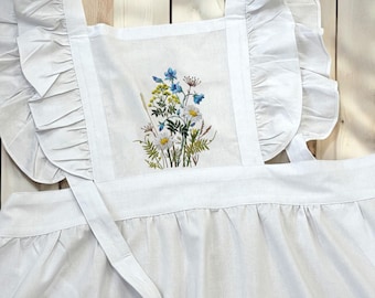 May include: White apron with a ruffled neckline and a floral embroidery design. The embroidery features blue and white flowers and green leaves.