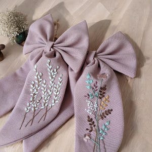 Puede incluir: Dos lazos de tela rosa polvoriento con dise&ntilde;os florales bordados. Un lazo presenta ramitas blancas de flores y el otro un ramo de flores multicolores.