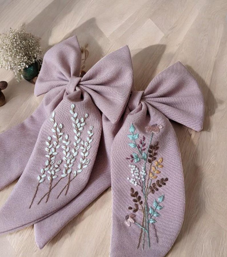 Puede incluir: Dos lazos de tela rosa polvoriento con dise&ntilde;os florales bordados. Un lazo presenta ramitas blancas de flores y el otro un ramo de flores multicolores.