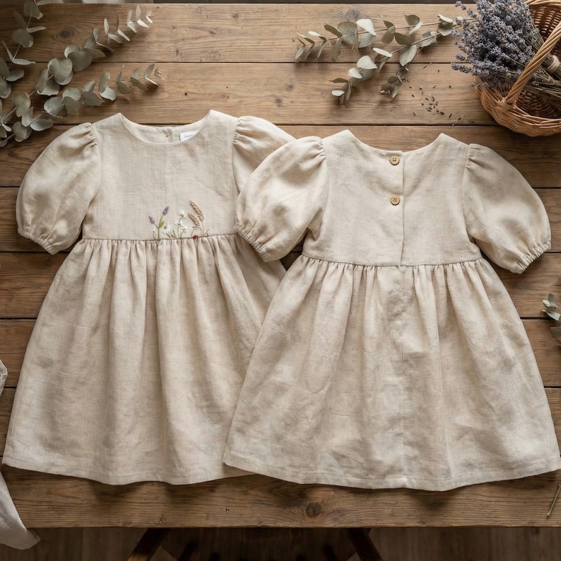 May include: Two beige linen dresses with puff sleeves and a gathered waist. The left dress features delicate floral embroidery on the chest. The right dress displays two small wooden buttons on the back closure. Both garments have a simple, classic cut.