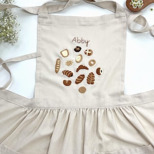 Puede incluir: Delantal beige con diseños de pan bordados y el nombre "Abby". El delantal tiene un peto, bolsillos en la cintura y lazos. El pan bordado incluye cruasanes, bagels y hogazas. El delantal está hecho de tela clara.