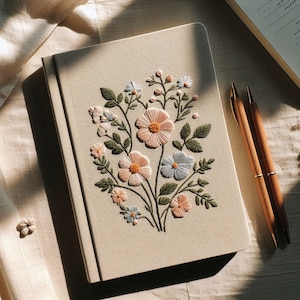 Op de afbeelding: Een beige notitieboek met een delicaat geborduurd bloemenmotief met roze en witte bloemen. De borduursels zijn gedetailleerd en ingewikkeld, wat een mooie en vrouwelijke esthetiek cre&euml;ert.