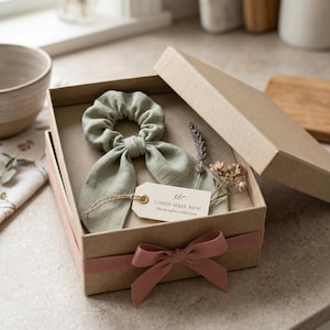 May include: A sage green linen hair bow scrunchie with long tails inside a kraft paper gift box. The box is tied with a dusty pink ribbon and includes a small tag that reads LINEN HAIR BOW Handcrafted with Care, alongside a sprig of dried lavender.