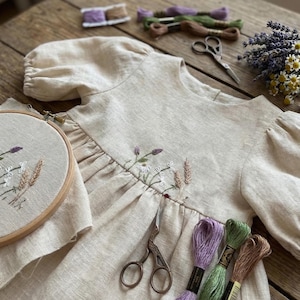 May include: A beige linen dress with puff sleeves featuring delicate floral embroidery across the bodice. An embroidery hoop with matching floral stitching, metal scissors, and several skeins of embroidery thread in purple, green, and brown are present.