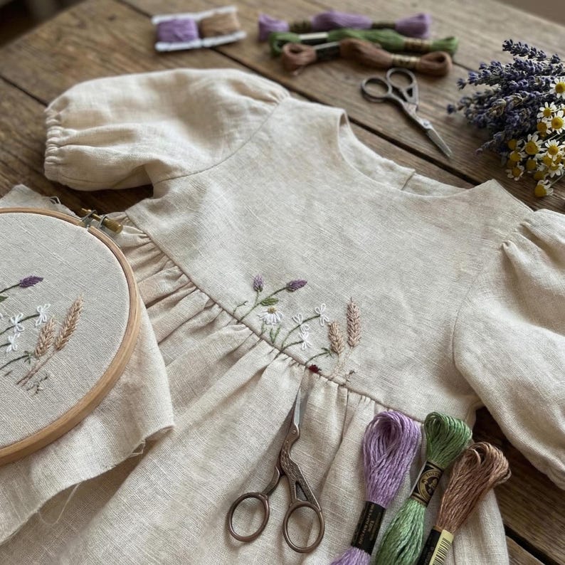 May include: A beige linen dress with puff sleeves featuring delicate floral embroidery across the bodice. An embroidery hoop with matching floral stitching, metal scissors, and several skeins of embroidery thread in purple, green, and brown are present.